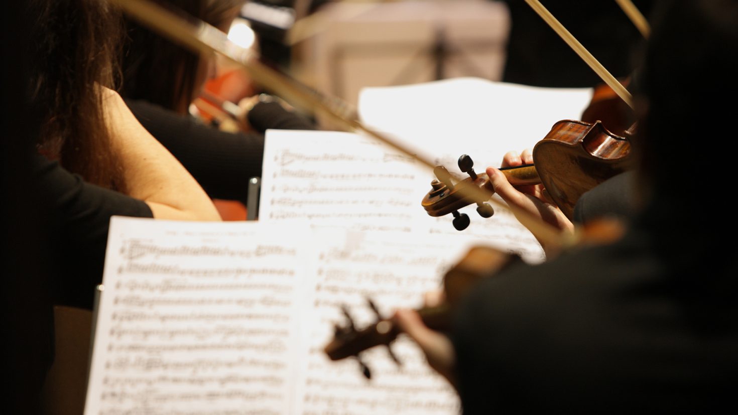 Orchestra musicians seen from the side, focusing on sheet music and string instruments such as violins. The image captures the hands of the musicians playing their instruments, with the sheet music visible in front of them. The background is softly blurred.