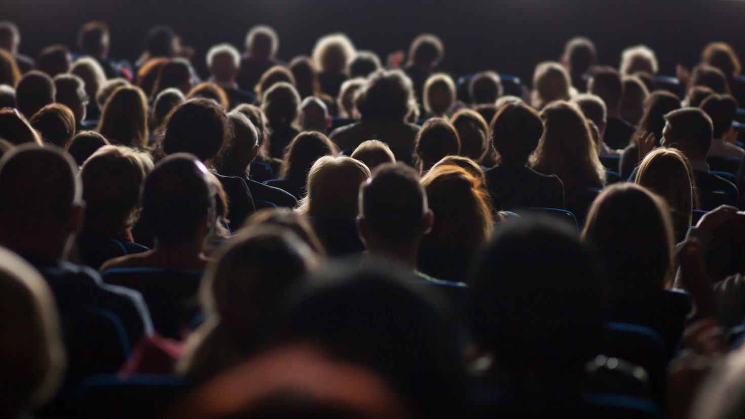 A large audience sits in a dimly lit theatre, focused on a presentation or performance happening off-screen. The image captures the backs of their heads, with illumination from the stage lights illuminating the tops of some individuals' heads.