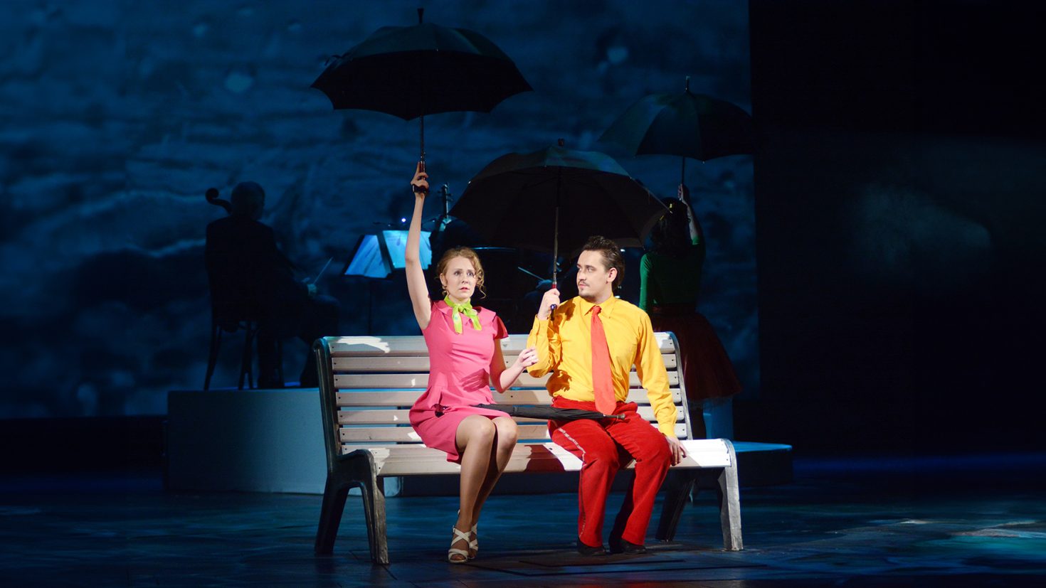 A woman and a man sit on a white bench, each holding a black umbrella. The woman wears a pink dress with a lime green bow, and the man wears a yellow shirt with a red tie and red pants. They are on a stage with a dark background and a musician playing in the distance.