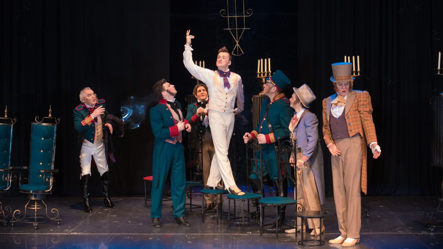 A theatrical scene with six actors on stage, dressed in elaborate period costumes. One actor in a white suit is standing elevated, striking a dramatic pose, while the others around him gesture expressively. Ornate chairs and candelabras serve as the background props.