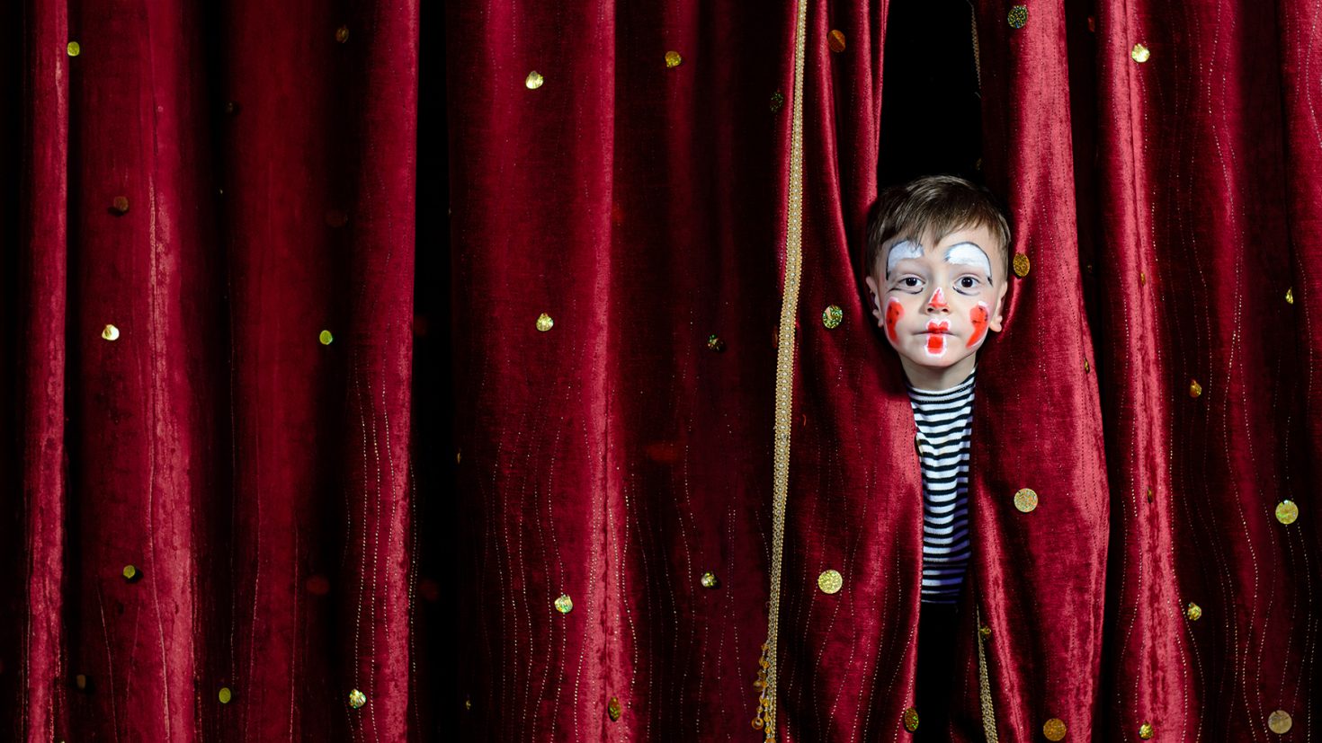 A young child with clown face paint and a striped shirt peeks through a gap in red, sparkly stage curtains adorned with gold dots.